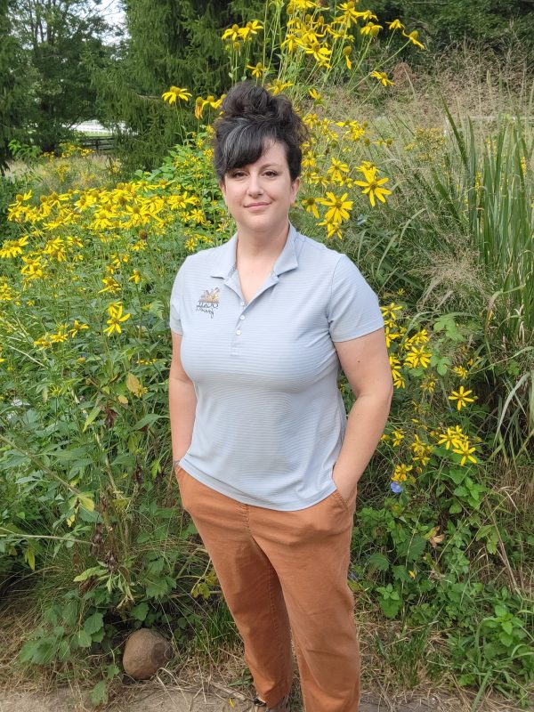 photo of woman with dark hair standing in front of flowering bush with yellow flowers wearing a gray polo shirt