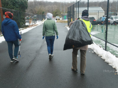 photo of the backs of three volunteers during the MLK Day of Service cleanup walking down a road in Barcroft park ringed with snow holding a trash picker upper and a large black trash bag. Photo credit YM Bird.