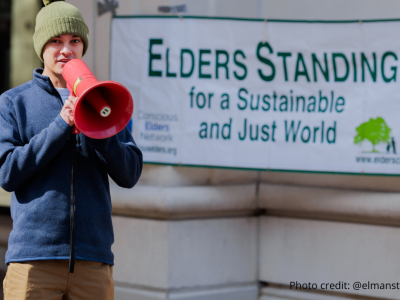 2025-Climate resolution-4 Juan Bustamante wearing a green wool ca and blue fleece, speaking to the crowd through a red megaphone. Photo credit: @elmanstudio