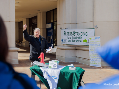 2025-Climate Resolution-5 Jim Lyon, EcoAction Arlington board member, holding up right fist and holding papers in left hand speaking in front of the crowd. There is a table with the Ecoaction Arlington branded green and white tablecloth, and red megaphone on table. In the background is the Bozman Government Center building, Elders Standing for a Sustainable and Just world banner sign, and an EcoAction Arlington sign. in the foreground are closeups over the shoulders of two rally-goers.