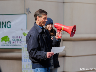 2025- Climate Resolution-7 John Bloom of Sierra Club Potomac River Group holding a red megaphone and an piece of paper, smiling and addressing the crowd. Molly Bolan is seen in the background. Photo credit: @elmanstudio.