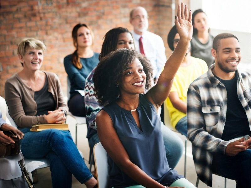 photo of diverse group of people casually dressed sitting in chairs with woman in foreground raising her hand