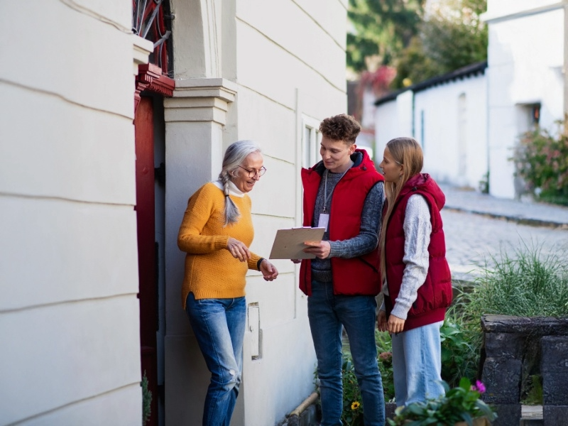 photo of two volunteers in red puffer vests with a clipboard speak to a home owner at her door