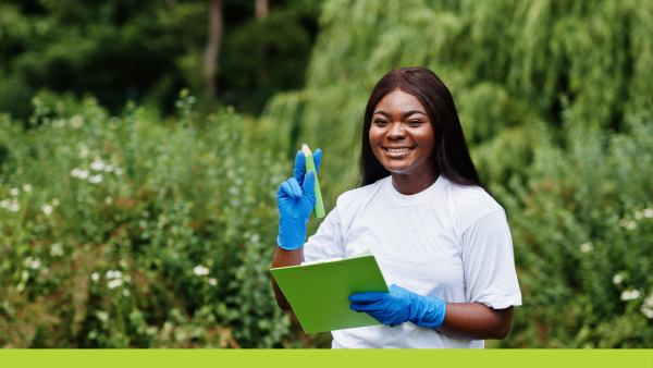 A smiling woman in a field wearing a white t-shirt and blue latex gloves holding a clipboard and giant pencil