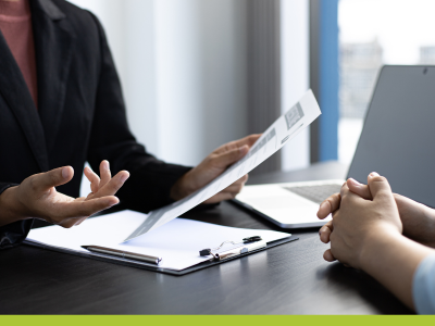 image of woman's torso in dark blazer holding a document in her hand and indicating with the other hand at a table with a clipboard and laptop and the folded hands of another person opposite