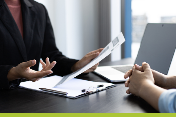 image of woman's torso in dark blazer holding a document in her hand and indicating with the other hand at a table with a clipboard and laptop and the folded hands of another person opposite