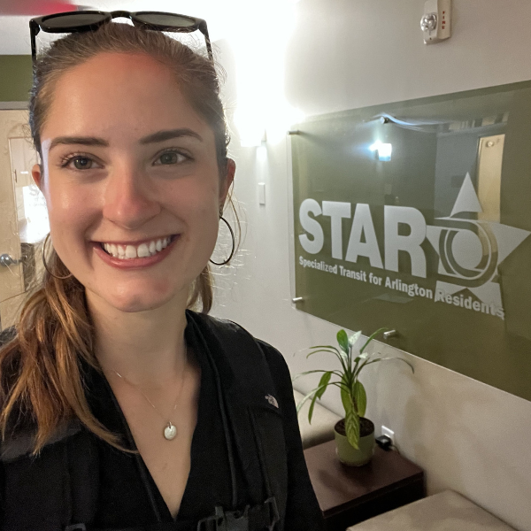 headshot of Lauren Breyer a smiling woman with long hair pulled back in a ponytail in a black blouse and sunglasses on top of her head, behind her is a sign that reads STAR Specialized Transit for Arlington Residents