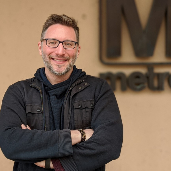 headshot of Mark Phillips a man with short brow hair and a beard wearing black-rimmed glasses, a black hoodie and black jacket with his arms crossed, behind him is a sign with a large M and metro.