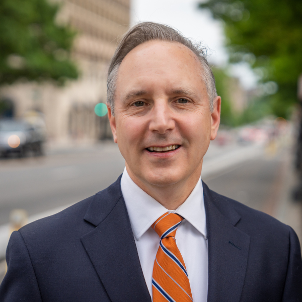 headshot of Stewart Schwartz a gentleman smiling and looking into the camera, wearing a navy suit, white shirt and red and blue striped tie