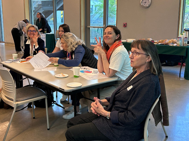 A group of females sitting around a conference table listening to a speaker. On of the group is looking at the camera smiling and making the peace sign