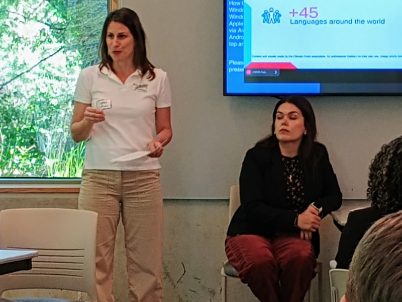 Crystal DiMiceli in a white polo and tan pants standing in front of a projection screen holding a sheet of paper in her left hand, to the side a woman is sitting in a chair under the screen