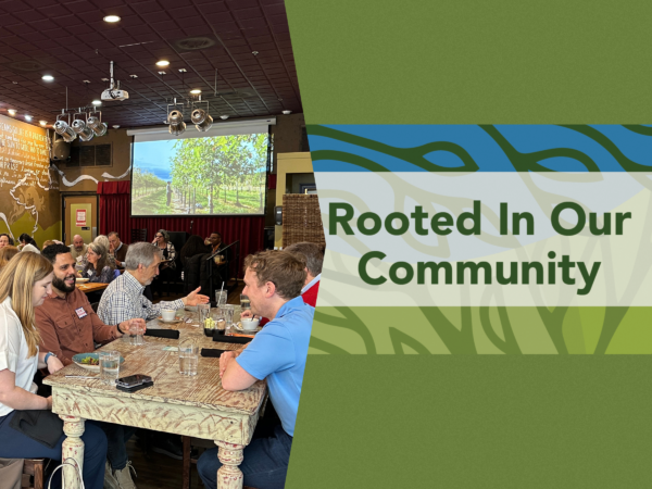 banner graphic with dark green background for a luncheon at Busboys & Poets Shirlington on Thursday, February 11, 2027 on the right is green and blue abstract leaf-like background with the bold text “Rooted In Our Community” centered across the image. On the right a photo of people sitting at tables with plates and glasses in front of them, with a colorful illuminated screen in the background depicting trees.